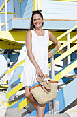 Brünette Frau in weißem Sommerkleid mit Flipflops und Korbtasche am Strand
