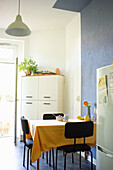 Table with yellow gingham tablecloth, black chairs, and white cupboard in kitchen with a high ceiling