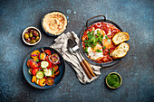 Shakshouka in der Pfanne mit Röstbrot, Gemüsesalat, Hummus und Oliven (traditionelles Frühstück oder Brunch aus dem Nahen Osten)
