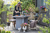 Frau dekoriert Gartentisch mit Osterdekoration, Kuchen, Osternest mit Eiern und Blumentöpfen