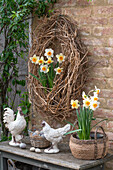 Clematiskranz an Steinmauer hängend, Narzissen (Narcissus) im Topf, Eier im Korb und Hühnerfiguren als Osterdeko