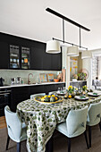 Langer, ovaler Esstisch und gepolsterte Stühle vor Küchenzeile mit dunklen Schrankfronten