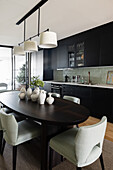Dunkler, ovaler Esstisch mit Keramiksammlung und Polsterstühle vor Küchenzeile mit schwarzen Schrankfronten