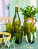 Grüne Gläser, schwarze Oliven in Schale, Olivenzweig, im Hintrgrund Krug mit Minze