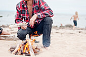 Mann kauert neben einem Lagerfeuer am Strand