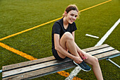 Smiling teenage girl tying shoelace while sitting on bench in playing field