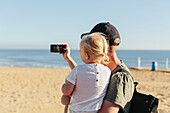 Vater trägt Tochter und macht ein Foto am Strand