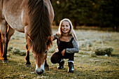 Smiling girl crouching on meadow with grazing pony
