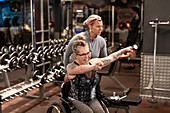 Glückliche Frau im Rollstuhl trainiert im Fitnessstudio mit Personal Trainer