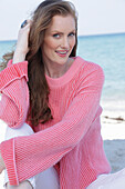 Junge, blonde Frau im rosafarbenen Pullover und weißer Hose am Meer