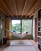 Narrow room with wood ceiling and a person lying in a hammock in front of window with garden view