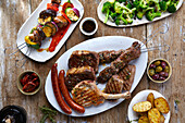 Grillplatte mit Würstchen, Schweinekotletts, Lammfleisch, daneben Gemüsespieße und Beilagen