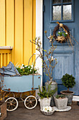 Vintage Kinderwagen mit Frühlingsblumen, alter Zinkkanister und Pflanzentöpfe vor der Eingangstür