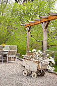 Vintage Kinderwagen mit Rhododendronzweigen auf Schotterplatz vor Gartentisch und Holzpergola auf der Terrasse