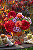Herbstblumenstrauß aus Dahlien (Dahlia) und Astern, Kerzen in Schalen von Esskastanien (Castanea Sativa) und Herbstlaub