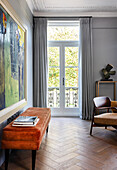 Orange-colored bench with velvet cover with artwork above it in formal living room with French doors