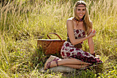 Blonde woman in off-the-shoulder summer dress with picnic basket on the meadow