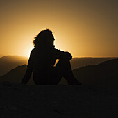 Silhouette einer sitzenden Frau mit Blick auf die Hügellandschaft des Valle De La Luna bei Sonnenuntergang; San Pedro De Atacama, Region Antofagasta, Chile