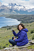 Eine Frau sitzt bei einem Getränk und einem Snack mit Blick auf einen See und eine zerklüftete Berglandschaft, Torres Del Paine Nationalpark; Torres Del Paine, Magallanes und Antartica Chilena Region, Chile
