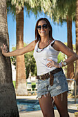 Eine junge Frau steht bei einer Palme und einem Swimmingpool; Palm Springs, Kalifornien, Vereinigte Staaten von Amerika