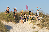 Eine Gruppe Jugendlicher rennt und springt über Sand und Strandgras; Tarifa, Cadiz, Andalusien, Spanien