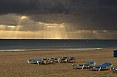 Sonnenstrahlen scheinen durch die dunklen Wolken über dem Meer und den Liegestühlen am Strand; Benidorm, Spanien