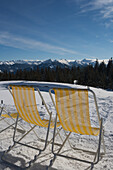 Ein Paar gelb-weiß gestreifte Liegestühle für Kunden im Sonnenschein vor einem Café am oberen Ende einer Skipiste mit Blick über die Berggipfel der österreichischen Alpen; Filzmoos, Österreich