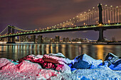 Manhattan Bridge By Snow-Covered Rocks At Sunset, Brooklyn Bridge Park; Brooklyn, New York, United States Of America
