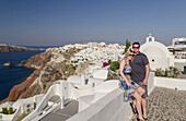 A Couple Posing Together On A White Wall On A Greek Island With The Town In The Background; Santorini, Greece