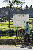 Female Cyclist Looking At A Large Map Sign Along A Canal With An Abbey In The Background; Saint-Gelven, Brittany, France