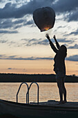 Silhouette eines Mädchens, das bei Sonnenuntergang eine brennende Papierlaterne in die Luft entlässt; Lake Of The Woods, Ontario, Kanada
