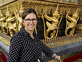 Portrait Of A Woman Standing Beside A Wall With Gold Statues, Temple Of The Emerald Buddha (Wat Phra Kaew); Bangkok, Thailand