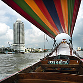 Boat With Colourful Covering And Buildings Along The Shoreline; Bangkok, Thailand