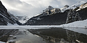 Canadian Rocky Mountains With Snow Under A Cloudy Sky Reflect In The Lake; Lake Louise, Alberta, Canada
