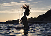 Silhouette einer Frau im hüfttiefen Wasser, die ihr nasses langes Haar in die Luft wirft; Tarifa, Cadiz, Andalusien, Spanien