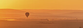 A Hot Air Balloon Flies Over The African Savannah In The Orange Light Before Sunrise, With A Trail Of Dust Below Made By A Truck On The Ground; Narok, Kenya