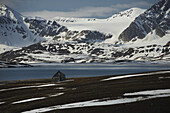 Kleine Holzhütte am Ufer des Arktischen Ozeans und schneebedeckte Berge; Spitzbergen, Svalbard, Norwegen