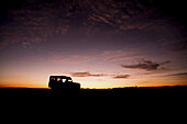 Ein auf der Savanne geparkter Lastwagen mit eingeschalteten Scheinwerfern ist als Silhouette am Horizont bei Sonnenaufgang zu sehen, blauer und oranger Himmel mit ein paar dünnen Wolken im Hintergrund; Narok, Kenia