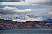 Hills Along The Coastline And Peaked Snow Covered Mountains Under A Cloudy Sky; Argyll, Scotland