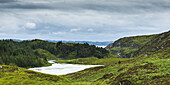 Ein Fluss, der in einen Teich fließt, umgeben von üppigem Gras und Wald in den Highlands; Schottland