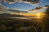 Die Sonne geht über dem Hafen von Ushuaia in Argentinien auf, als ein Schiff in die ruhige Bucht einläuft, mit Bäumen im Vordergrund und niedrigen Hügeln am Horizont, während der Himmel mit Wolken übersät ist und sich von einem goldenen Gelb zu einem tiefen Blau verändert; Ushuaia, Argentinien
