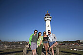 A Group Of Friends Standing With A Lighthouse In The Background Outside Of Amsterdam Central; Egmond Aan Zee, Holland