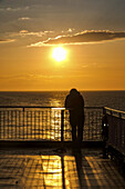 An Adult Stands At The Railing Looking Out At The North Sea With A Golden Sunset; England