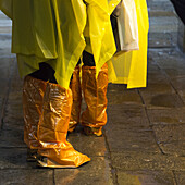 Stehende Menschen mit gelben Regenponchos und Schuhen, die mit orangefarbenen Plastikplanen bedeckt sind; Venedig, Italien