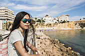 A Chinese Young Woman In Sunglasses Stands At A Railing Looking Out At The Water; Benidorm, Alicante, Spain