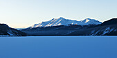 Frozen Snow Covered Lake With Snow Covered Mountains In The Background, Liard River Hot Springs Provincial Park; British Columbia, Canada
