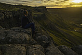 Person auf einer Klippe sitzend mit Blick auf den Sonnenuntergang in der Nähe der Stadt Djupavik; Island