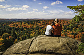 Paar sitzt auf einer Klippe mit Blick auf die Herbstfarben im Algonquin Park, Frau hat Kopf auf der Schulter des Mannes; Ontario, Kanada
