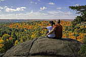 Junges Paar auf einer Klippe sitzend mit Blick auf die Herbstfarben des Algonquin Park; Ontario, Kanada