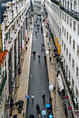 Fußgänger mit Regenschirmen gehen die schmale Straße zwischen den Gebäuden entlang; Lissabon, Region Lisboa, Portugal